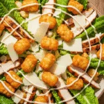 An overhead view of a large bowl of crispy tater caesar salad with golden smashed tater tot croutons, chicken, and parmesan cheese.
